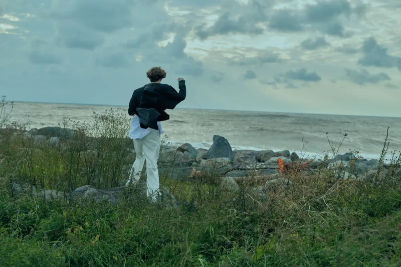 A lone walker stands on a rocky shoreline, facing the sea under a cloudy sky, jacket and white pants catching the breeze, capturing a calm outdoor seascape and rugged coast mood