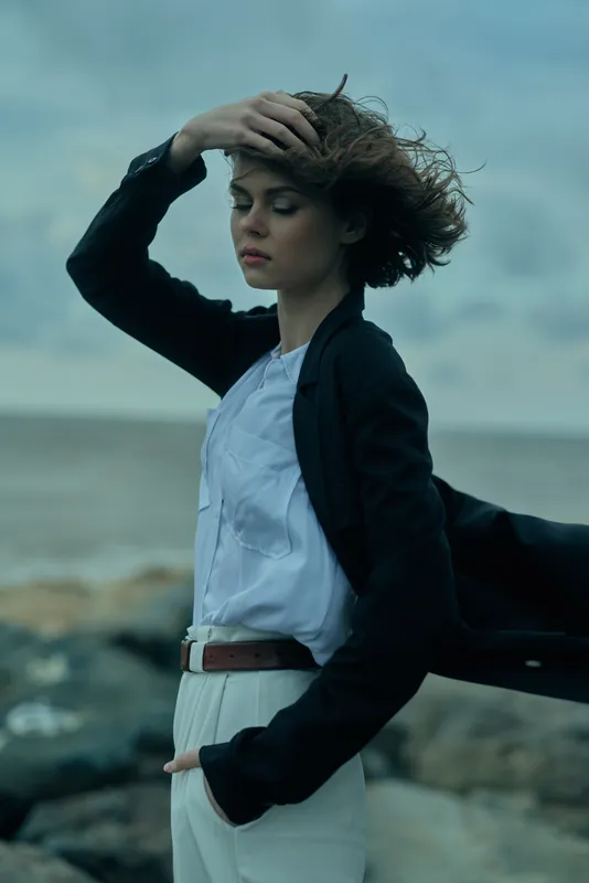 Woman stands by a windy coastline in a blazer and white shirt, hair swept by the breeze, conveying confident fashion and coastal style with open sea and rocky background
