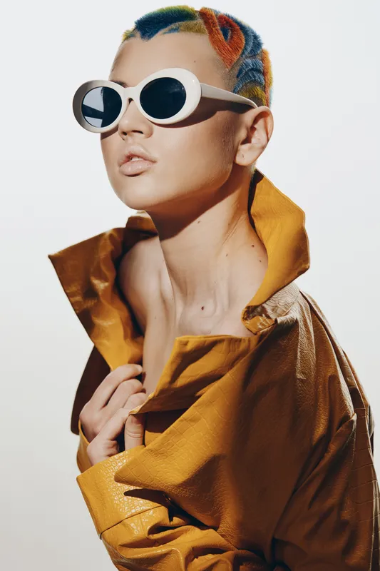 Young woman with colorful braided hair and white sunglasses wearing a mustard leather jacket, striking a confident pose against a bright neutral background, fashion portrait.