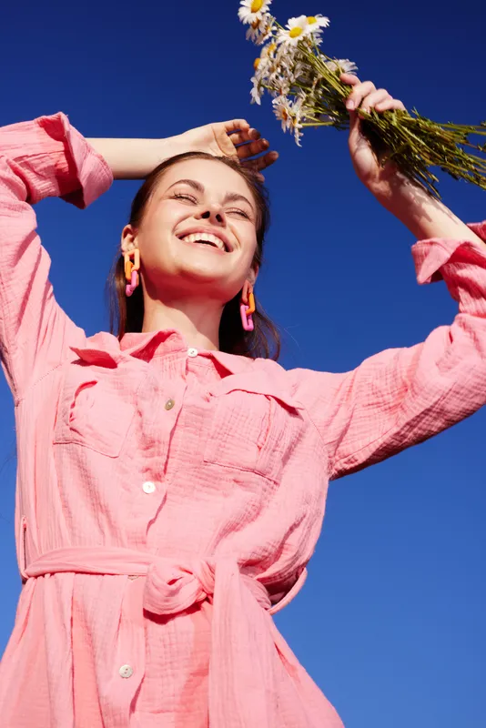 Joyful young woman with flowers against a blue sky, wearing a pink shirt dress, embodying happiness and summer vibes in a vibrant outdoor setting