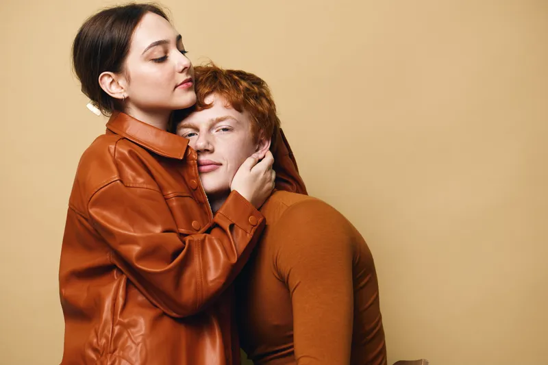Couple embracing with closed eyes in warm brown and orange clothing against beige background showing affection and calm connection between young people in studio.