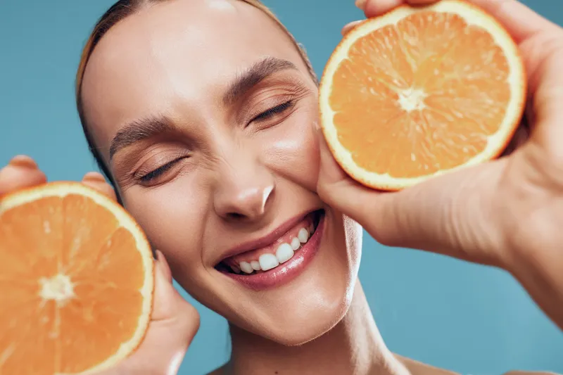 Happy woman holding two orange halves close to her face smiling with eyes closed against blue background, fresh fruit concept, healthy lifestyle, natural light.
