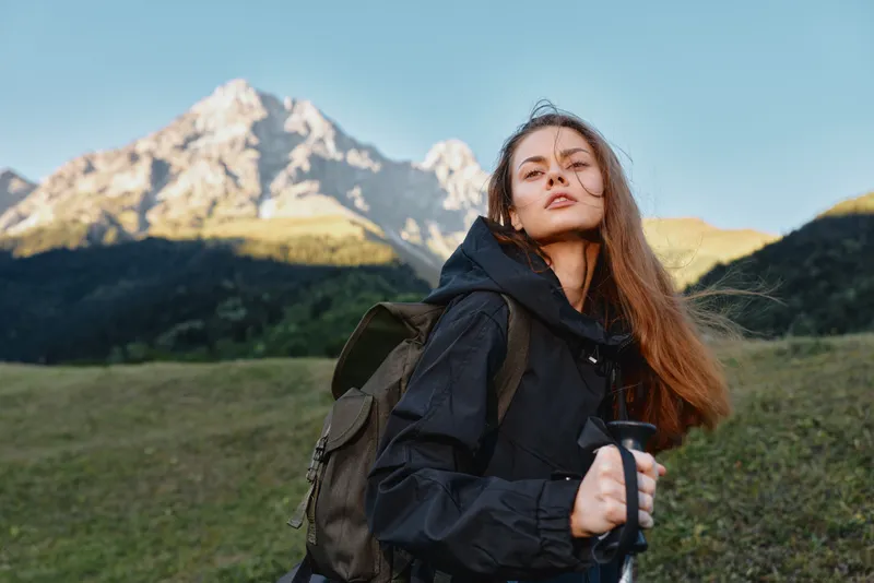 A young woman stands outdoors in a mountain landscape, wearing a dark jacket and backpack, looking toward the camera with a confident, adventurous expression.