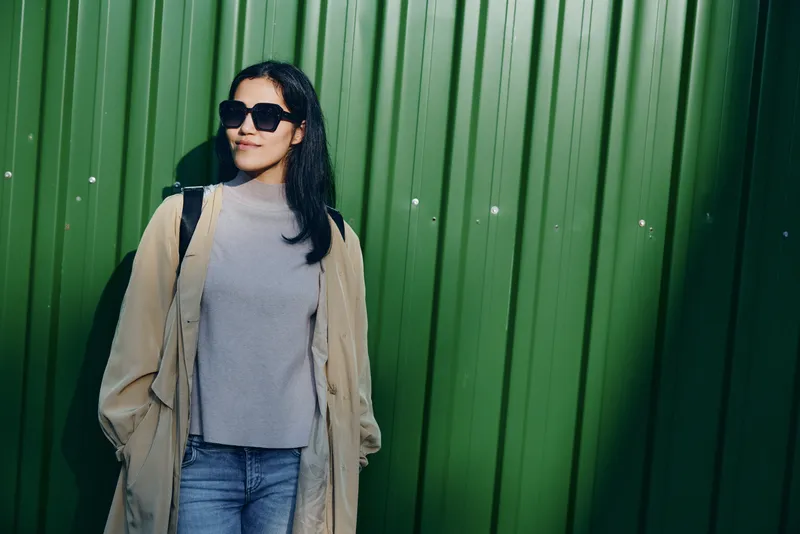 Woman wearing sunglasses, beige coat and jeans stands by a green wall on a sunny day, showcasing casual street fashion with a confident urban vibe and relaxed pose.