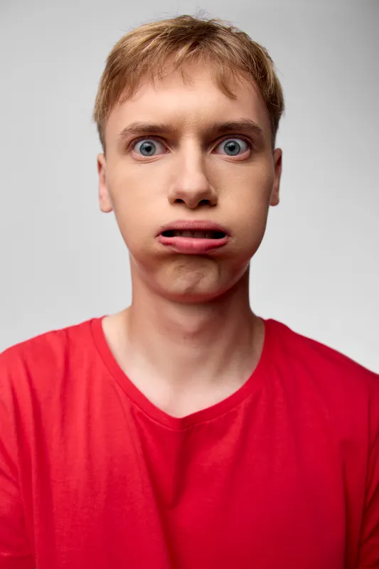 Awkward expression, confused face, young man, front view, red shirt, surprised look, facial expression, uneasy mood, closeup shot of a tense, candid moment under studio lighting