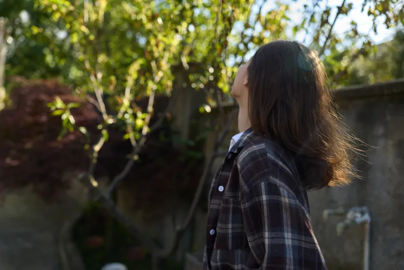 Casual woman outdoors, relaxed lifestyle moment, plaid shirt, sunlit garden setting, hair in breeze, thoughtful pose, natural colors, lush greenery background, candid portrait showcasing confidence