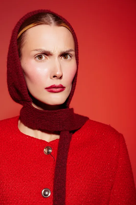 Portrait of a serious woman wearing a red scarf and coat against a red background. She has a focused expression with bold red lipstick and smooth skin. Fashion concept.