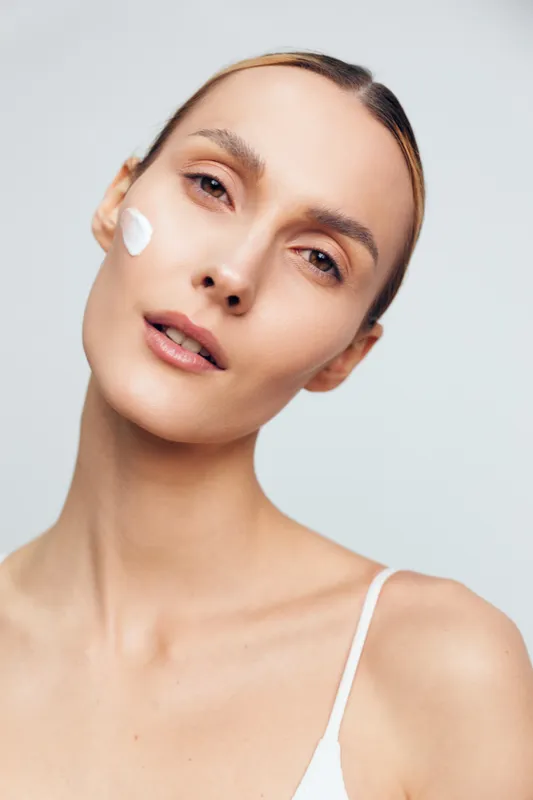 A young woman with clear skin applies white cream on her cheek, looking directly at the camera. The minimal makeup and light background emphasize skincare and natural beauty.