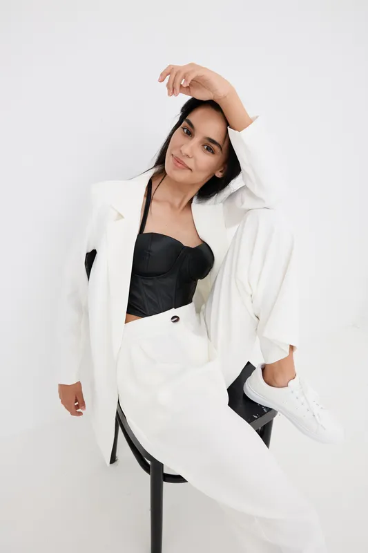 stylish woman wearing white suit and black corset seated on chair with relaxed pose and confident expression, minimalistic white background in bright studio lighting
