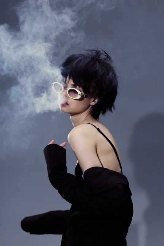 Stylish woman smoking with sunglasses in moody dark setting, exhaling smoke with edgy fashion and modern hairstyle.