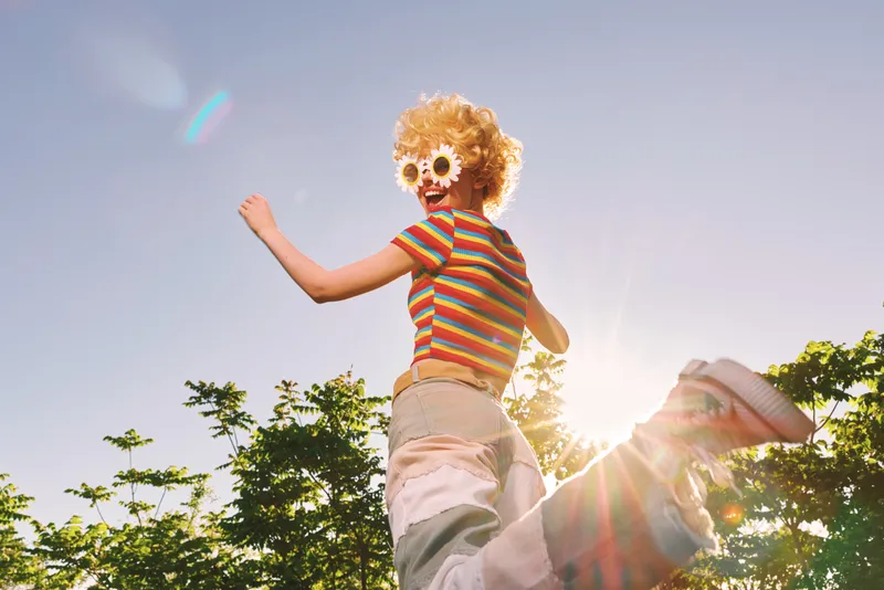 Playful woman outdoor scene with retro vibes, striped shirt, oversized sunglasses, curly wig, and carefree mood, jumping above the camera as sun flares, bold colors, candid lifestyle moment.