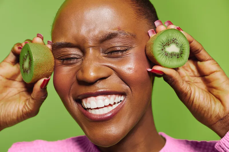 Smiling black woman with dark skin holds two halves of fresh kiwi fruit near her face against green background. Concept of healthy eating, natural food, and joyful lifestyle with tropical fruit.