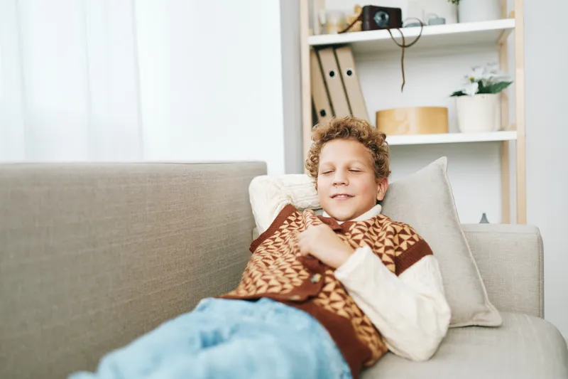 Young boy rests comfortably on a sofa, eyes closed, cozy blanket wrapped warm over shoulders, enjoying a peaceful moment in a bright living room with soft light and calm ambience.