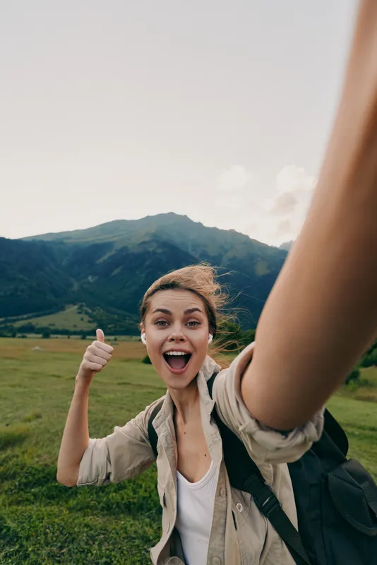 A happy woman in casual clothes takes a selfie outdoors with mountains in the background, giving a thumbs up. Concept for lifestyle, travel, and adventure.