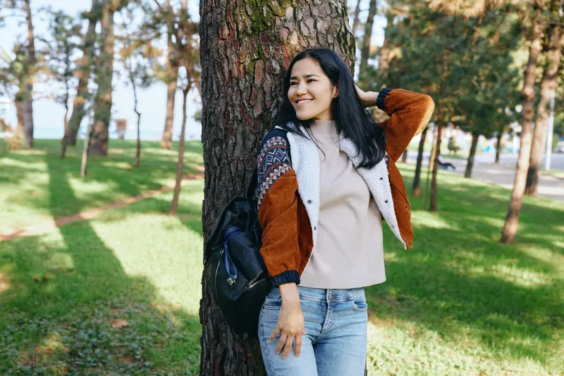 Woman, tree, park, outfit, casual, smile, sunlight, nature, relaxation, outdoor scene of a woman leaning against a tree in a sunny park, wearing a beige sweater, colorful jacket and jeans, enjoying a