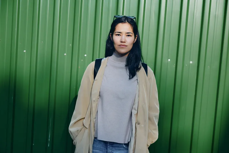 Woman in a casual beige coat and gray turtleneck stands before a green wall, urban fashion mood, relaxed street style and everyday wear, candid photograph.
