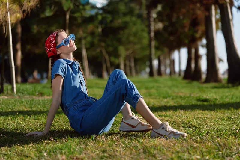 Lifestyle woman in modern boho-western denim street style relaxing outdoors on grass with film color tones, wearing sunglasses and a red bandana under natural sunlight.