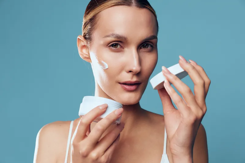 Woman applies skincare cream to face, beauty hydration concept, blue studio background, cotton pads in hands, gel texture on cheeks, calm look, cosmetic treatment, spa hygiene