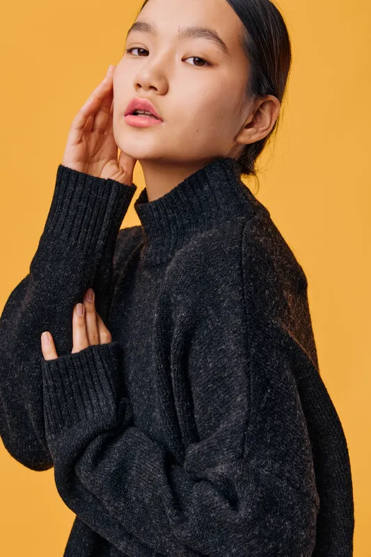 Young woman with natural makeup in black sweater posing on yellow background with thoughtful expression, fashion portrait of female model with dark hair and casual style clothing.