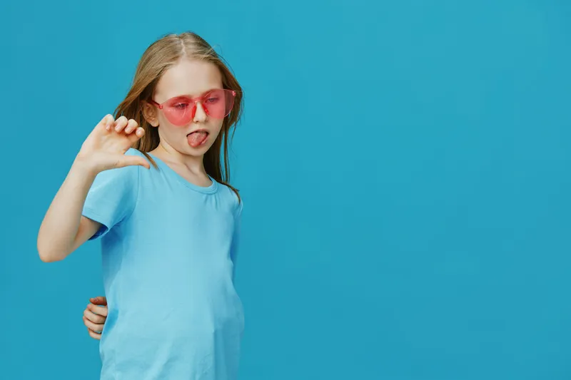 Playful girl in pink sunglasses making a funny face against a vibrant blue background, showcasing youthful energy and summer vibes