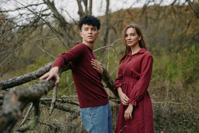 Two individuals stand in a rustic field, showcasing authenticity through relaxed pose, natural expression, and genuine connection beneath autumnal trees and muted light.