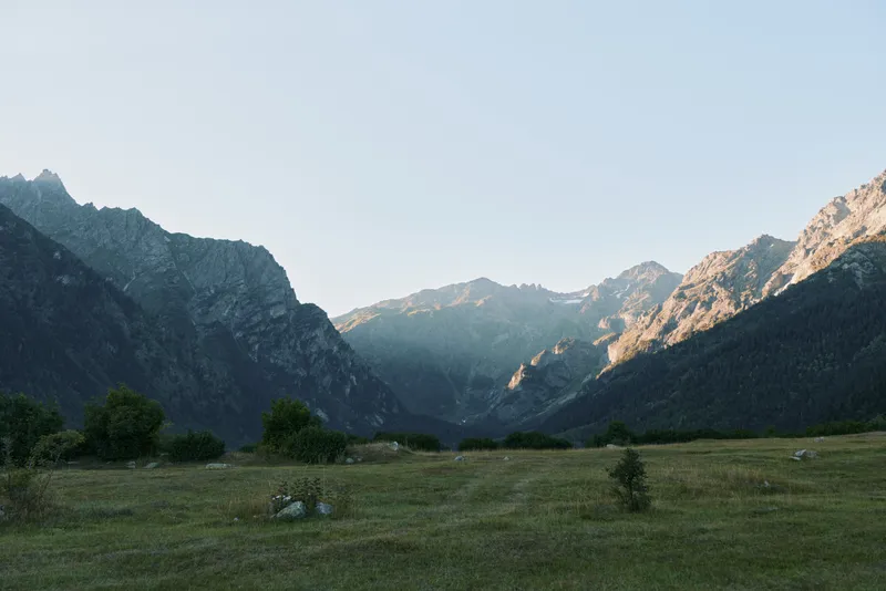 A tranquil alpine valley unfolds beneath rugged peaks as warm sunlight highlights a vast meadow, creating a serene natural landscape ideal for outdoor exploration and peaceful reflection.