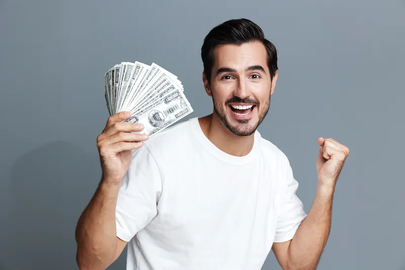 Happy smiling man holding cash money with excited expression celebrating success isolated on grey background. Financial achievement concept.