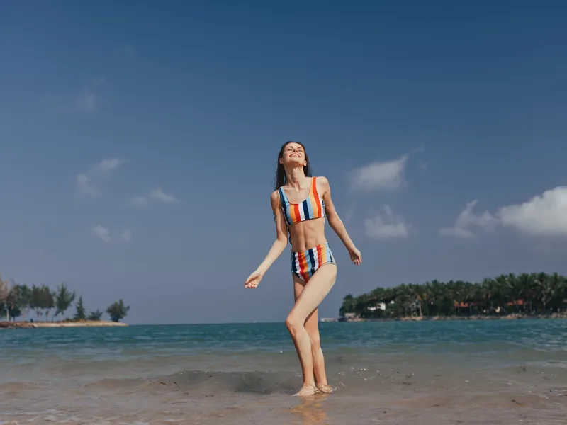 A young woman wearing a striped bikini stands barefoot on the shoreline, smiling as waves lap her feet under a clear blue sky, conveying carefree summer joy and freedom.