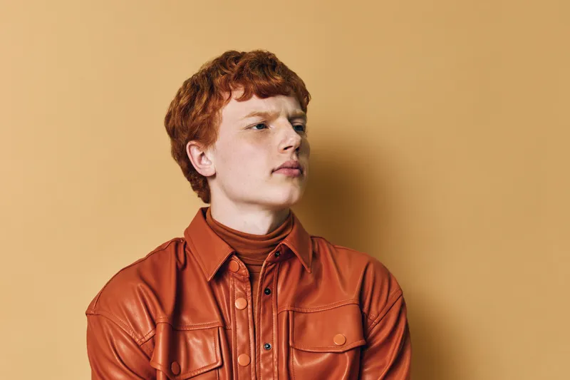Young man with red hair and curly hairstyle wearing orange leather jacket looking sideways against beige background. Portrait of confident thoughtful teenager with casual fashion style.