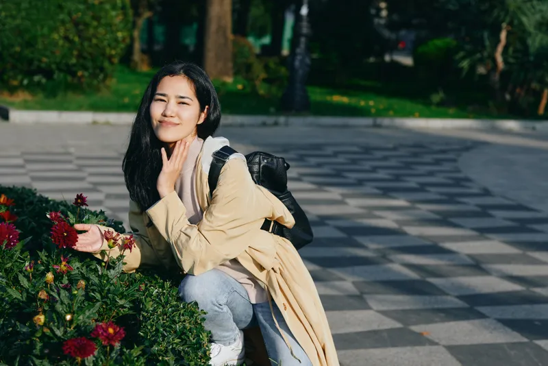 Woman crouching beside a flower bed in a park, wearing a light coat and backpack, enjoying a sunny day among blooming plants and greenery, relaxed pose and gentle smile
