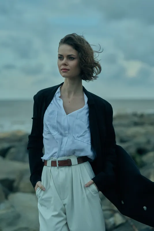 Woman standing on a rocky coastal shore, wind in hair, wearing a blazer over a white blouse and high waisted trousers with a brown belt, looking toward the horizon in a moody sea breeze setting.