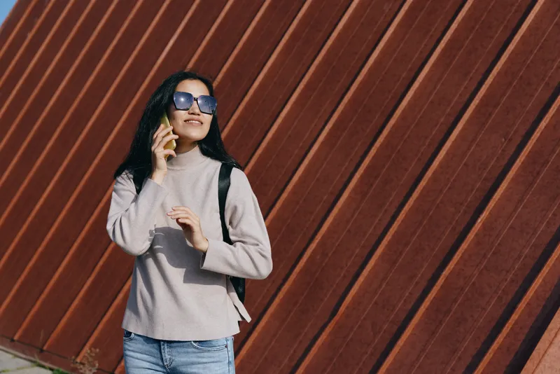 Woman on phone outdoors, wearing sunglasses, sweater and jeans with a backpack, standing beside a diagonal timber wall for a casual urban lifestyle moment