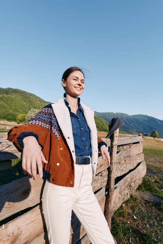 woman countryside fence mountains jacket smiling outdoors portrait. Young woman stands leaning on a wooden fence in a rural landscape wearing a patterned jacket, smiling confidently under clear blue