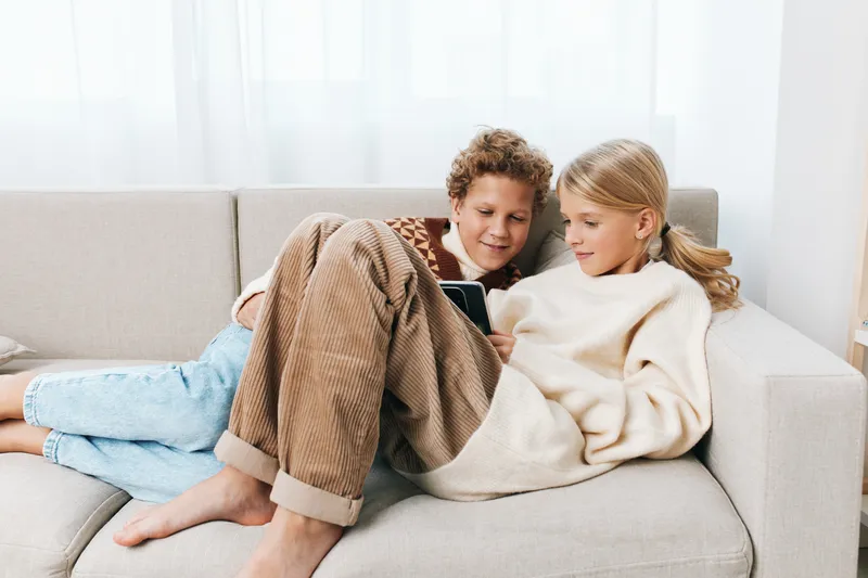 Authenticity shines as siblings share a cozy moment on a couch, exploring a tablet together, capturing a playful school lifestyle scene in a warm home, and quiet atmosphere for viewers.