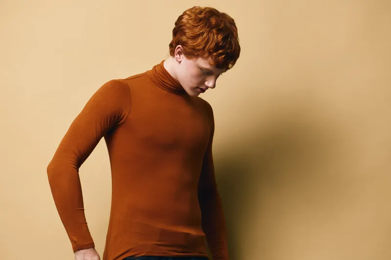 Young man with curly red hair wearing brown turtleneck sweater looking down with serious expression isolated on beige background in studio portrait.