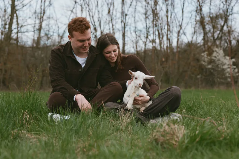 Couple sitting on the grass with a playful goat kid in a meadow, outdoors with trees in the background. Cozy, intimate moment highlighting companionship, happiness, and countryside lifestyle.
