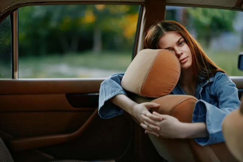Thoughtful young woman resting her head on a car seat, reflecting on life, dressed casually in denim, with a serene outdoor background.