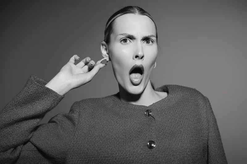 Surprised young caucasian woman with light skin pulls ear and opens mouth in shock, wearing textured sweater against grey background in studio portrait.