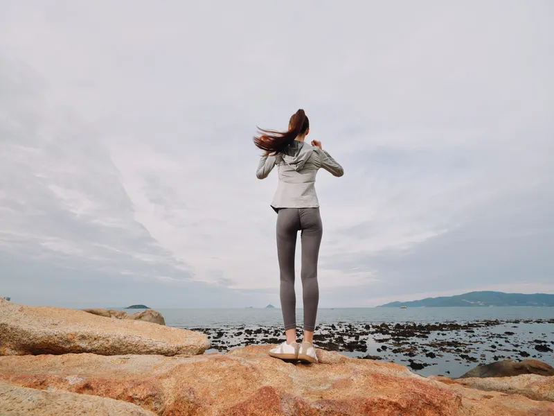 A young woman in activewear standing on rocks by the ocean, enjoying nature s tranquility and showcasing a healthy lifestyle in a serene coastal setting