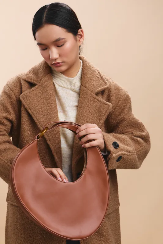 Asian woman in a warm coat inspects a stylish circular brown handbag, standing in a minimalist studio with soft lighting and a calm, focused mood.