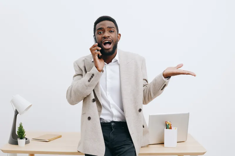 Confused man talking on mobile phone in office workspace with laptop and plant, expressing surprise or frustration, casual business attire, light background, modern office environment, workplace commu