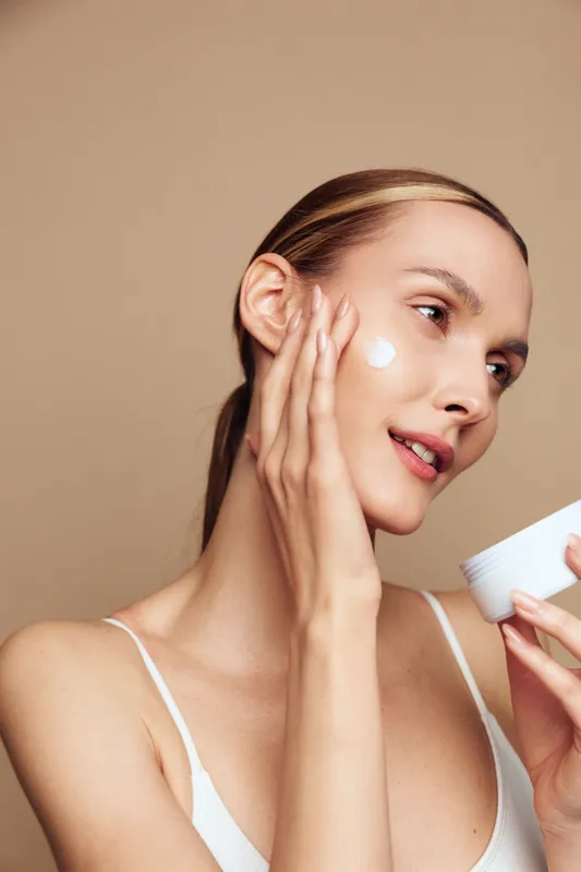 A young woman with clear skin applies face cream on her cheek using her fingers in a softly lit studio. Concept for skincare, beauty routine, and natural skin health.