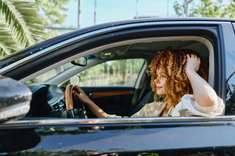 woman driving car with curly hair on sunny day, casual clothing, relaxed expression, modern vehicle interior, natural light, outdoor setting