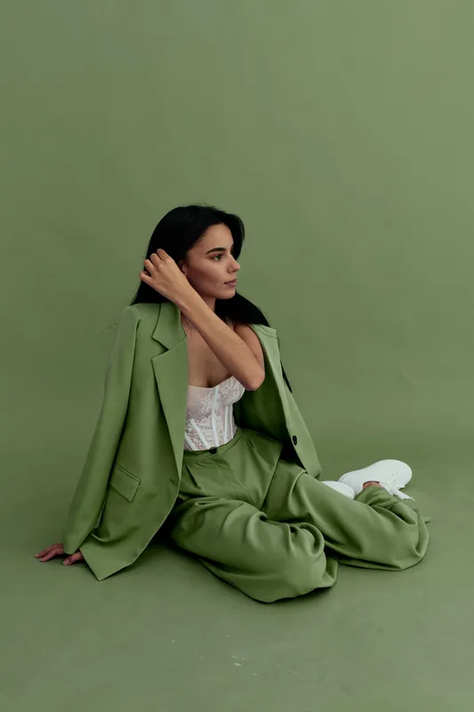 Young woman in stylish green suit sitting on the floor against a green background, wearing white lace top and sneakers, posing elegantly with calm expression.