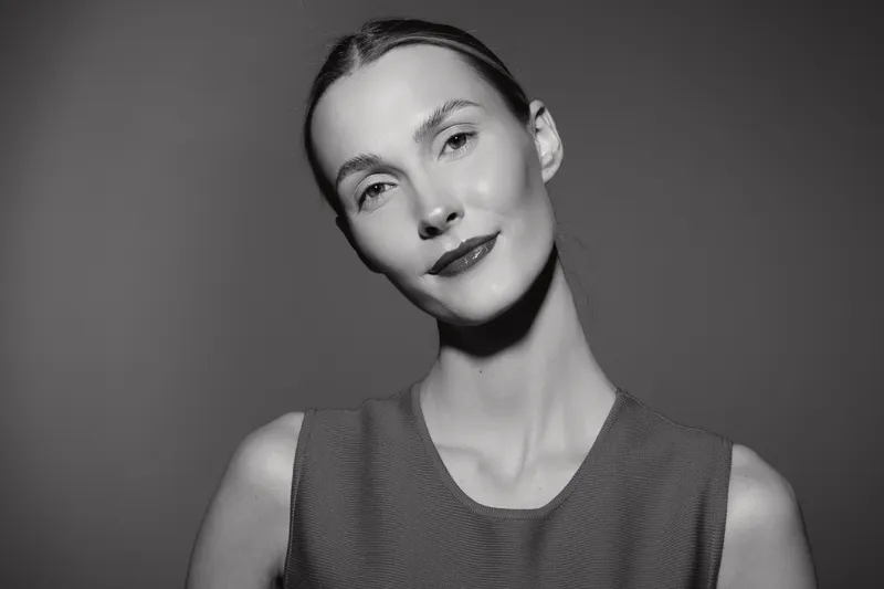 Portrait of a young white woman with light skin and slicked-back hair wearing a sleeveless top, looking confidently at the camera in black and white studio lighting.