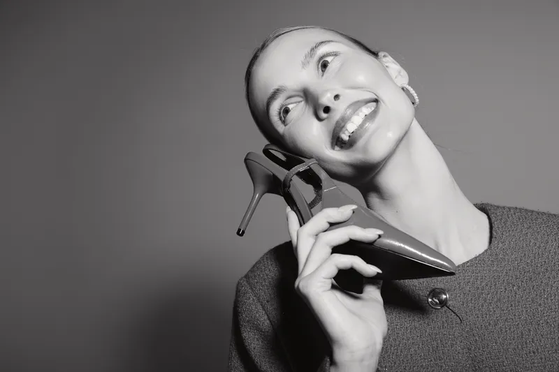 Smiling caucasian woman with fair skin holding high heel shoe near face in studio. Concept of fashion, elegance, happiness and style.