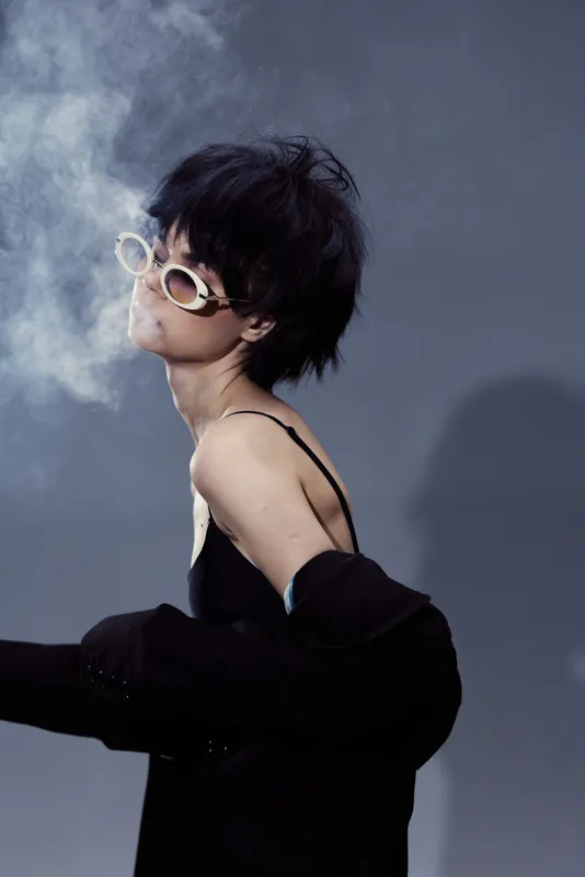 Stylish woman with short hair and vintage sunglasses blowing smoke in dark clothing against a grey background.