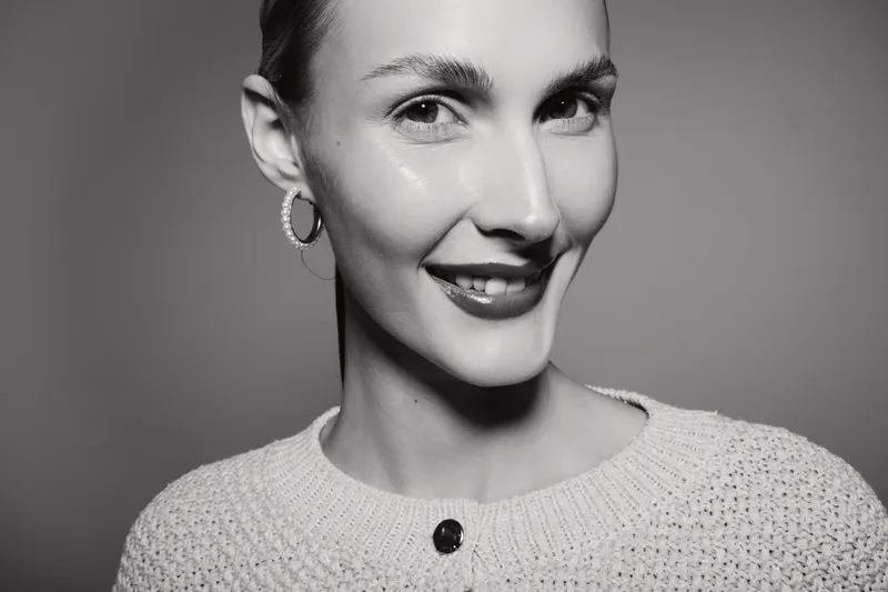 Smiling young Caucasian woman with light skin wearing knitted sweater and hoop earrings looks at camera in studio portrait.