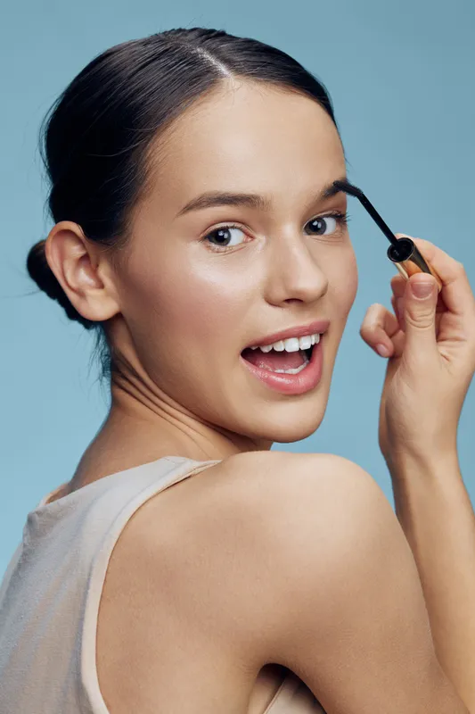 Young woman applying mascara in front of a soft blue background, showcasing beauty and confidence with a cheerful expression, perfect for beauty and makeup themes