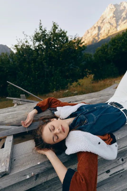 Woman portrait relaxing on wooden deck in nature with mountains in background, eyes closed and cozy layered denim and shearling jacket, peaceful outdoor leisure moment and soft evening light